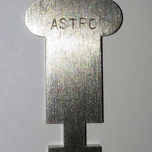 May include: A silver metal key with a unique shape and the word "ASTRO" engraved on the top. The key has a rectangular base and a long, narrow shaft.