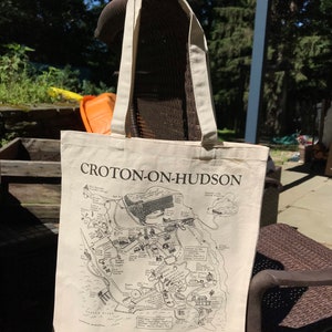 K&ouml;nnte beinhalten: Eine wei&szlig;e Canvas-Tragetasche mit schwarzen Henkeln und einer schwarz-wei&szlig;en Karte von Croton-on-Hudson, New York. Die Karte zeigt verschiedene Sehensw&uuml;rdigkeiten und Punkte von Interesse, darunter das Croton Reservoir, der Croton Aqueduct und der Croton Point Park.