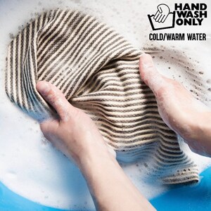 K&ouml;nnte beinhalten: Eine Hand w&auml;scht ein gestreiftes Stoffst&uuml;ck in einem Waschbecken mit Seifenwasser. Die Anweisungen "HAND WASH ONLY COLD/WARM WATER" sind auf einem wei&szlig;en Etikett aufgedruckt.