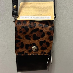 May include: A small, black and leopard print crossbody bag with a black leather strap. The bag has a flap closure with a silver button. The tag reads "STYLE A027283 COLOR: LEOPARD/BLKDIST FC: WGI."