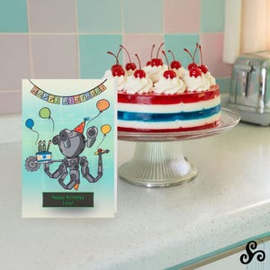 May include: A birthday card with a robot illustration holding a cake and balloons, with the text "Happy Birthday Lucy!" A layered red, white, and blue gelatin cake topped with whipped cream and cherries sits on a glass cake stand.