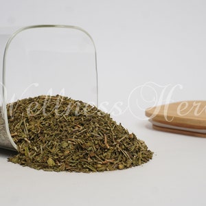 May include: A glass jar with a wooden lid filled with dried green herbs. The jar is tipped over and the herbs are spilling out onto a white surface.