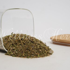 May include: A glass jar with a wooden lid, filled with dried green herbs. The jar is tipped over, spilling the herbs onto a white surface.