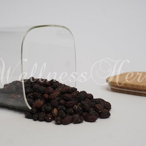 Puede incluir: Un tarro de vidrio con tapa de bambú, volcado, derramando bayas secas de color marrón oscuro. El tarro es transparente y tiene forma cuadrada. La tapa es de color marrón claro y tiene una superficie lisa.