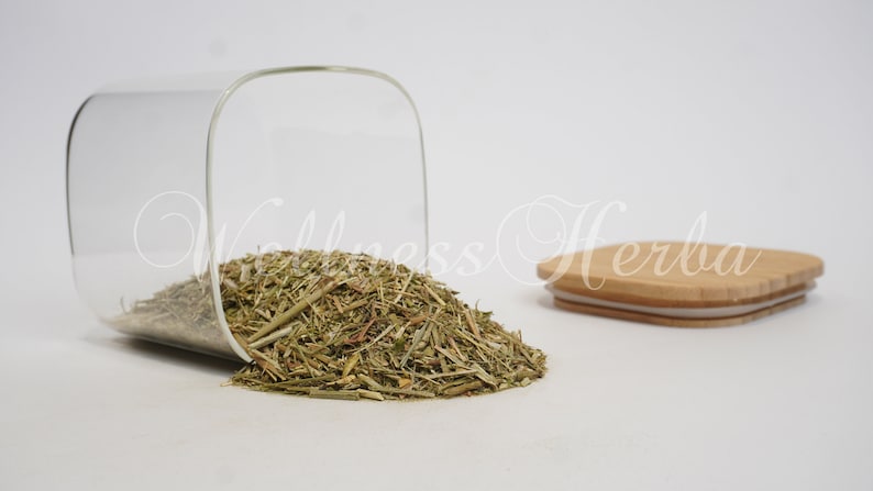 Puede incluir: Un tarro de vidrio con tapa de madera, lleno de hierbas verdes secas. El tarro est&aacute; volcado, derramando las hierbas sobre una superficie blanca.