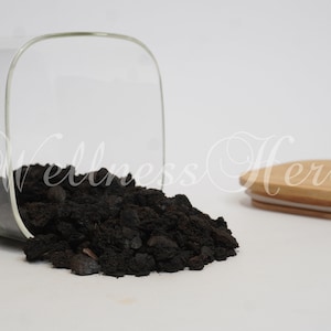 Puede incluir: Un tarro de vidrio con tapa de bambú, volcado, derramando roca volcánica negra. El tarro es transparente y la tapa es marrón claro.