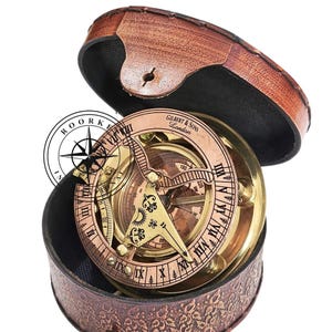 Puede incluir: Una brújula de latón con una funda de cuero. La brújula tiene un reloj de sol y una rosa de los vientos. La funda es marrón con un patrón floral. La brújula está etiquetada "Gilbert & Sons London".