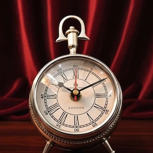 May include: A silver-colored, round clock with Roman numerals and black hands. The clock face reads "LONDON" and has a small handle at the top. The clock is set on a dark wooden surface against a red backdrop.