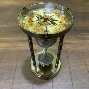 Puede incluir: Un reloj de arena antiguo de latón con una brújula en la parte superior. La brújula tiene un diseño detallado con marcas direccionales. El reloj de arena contiene arena oscura. La base y el marco tienen una apariencia envejecida.