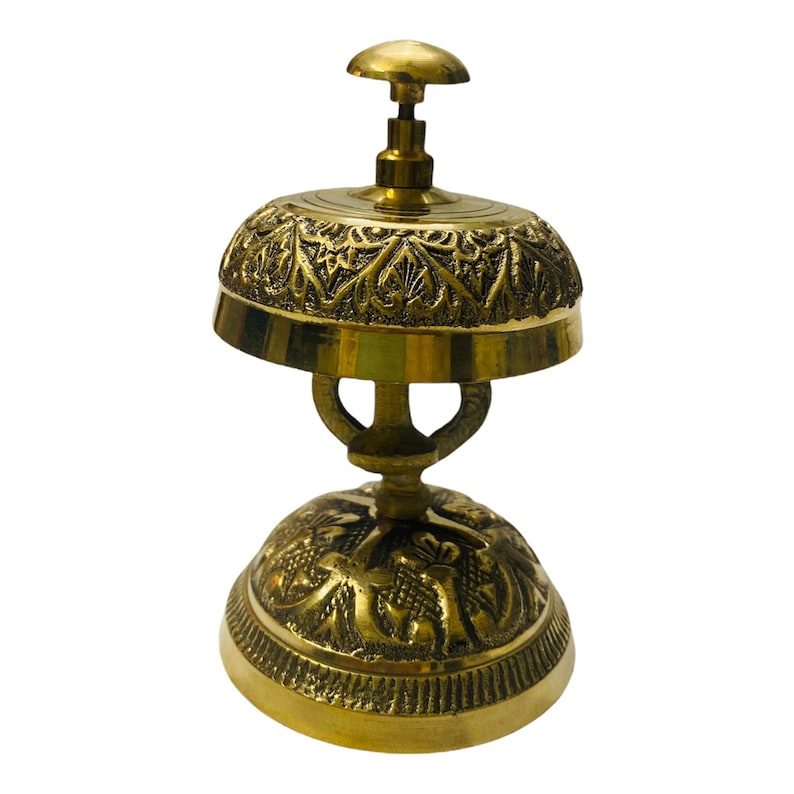 May include: A gold-toned brass hotel service bell with intricate floral designs. The bell has a round top and a base with a decorative pattern.
