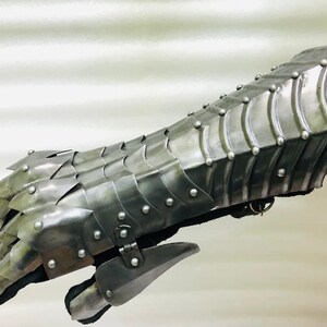 May include: A silver metal gauntlet with a scaly design and rivets. The gauntlet is made of metal plates that are connected by rivets. The gauntlet is designed to protect the hand and forearm.