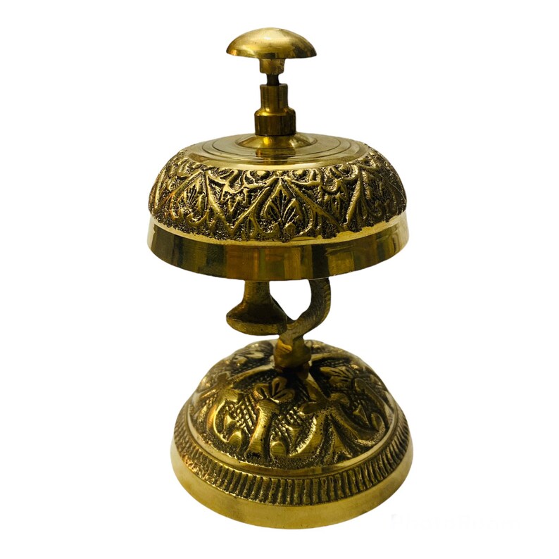 May include: A gold-toned brass hotel service bell with an ornate design. The bell has a rounded top and a base with intricate details.