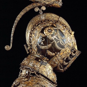 K&ouml;nnte beinhalten: Ein detaillierter, verzierter Helm aus Gold und Silber mit einer gefl&uuml;gelten Figur auf der Oberseite. Der Helm ist mit komplizierten Mustern und Designs verziert.
