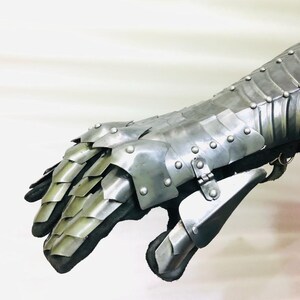 May include: A silver metal gauntlet with a segmented design and rivets. The gauntlet is made of multiple plates that are connected together. The gauntlet is designed to protect the hand and forearm.