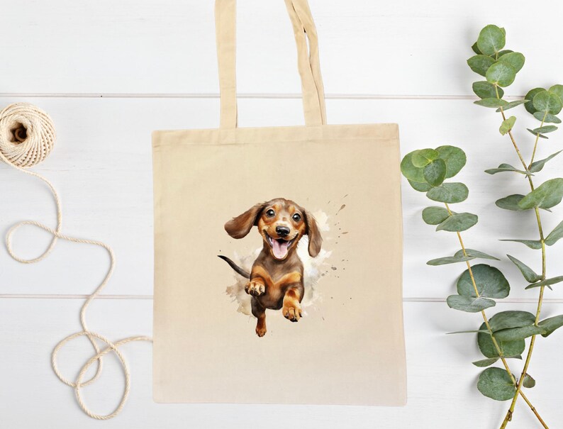 Peut inclure: Un sac fourre-tout en toile beige avec une illustration &agrave; l'aquarelle d'un chien teckel heureux. Le chien est marron et beige avec la langue sortie, semblant sauter hors d'une &eacute;claboussure de peinture. Le sac a deux anses beiges.