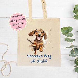 Peut inclure: Un sac fourre-tout en toile beige avec une illustration &agrave; l'aquarelle d'un chien teckel. Le chien est marron et beige avec une expression joyeuse. Le texte "Snoopy's Bag of Stuff" est imprim&eacute; en bleu. Le sac a des anses beiges.