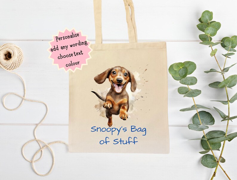 Peut inclure: Un sac fourre-tout en toile beige avec une illustration &agrave; l'aquarelle d'un chien teckel. Le chien est marron et beige avec une expression joyeuse. Le texte "Snoopy's Bag of Stuff" est imprim&eacute; en bleu. Le sac a des anses beiges.