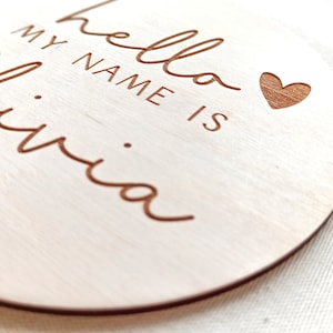 Op de afbeelding: Ronde houten hanger met de gegraveerde tekst "hello my name is Olivia" en een hartje. De hanger is gemaakt van lichtgekleurd hout met een natuurlijke nerf en een donkerbruine rand.