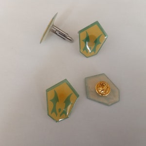May include: A set of four cufflinks and a pin featuring a green and yellow shield design. The shield has a stylized green creature inside.