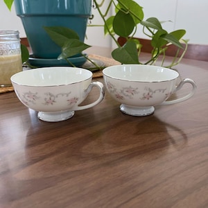 May include: Two white teacups with silver trim and floral designs. The teacups have a pedestal base and curved handles. The floral pattern includes pink flowers and green leaves. The teacups are placed on a wooden surface.