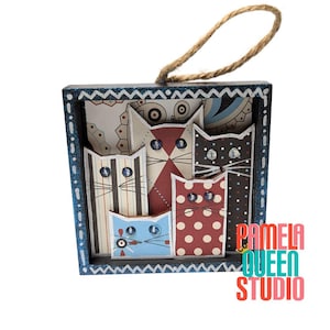 May include: A decorative wooden box featuring a family of stylized cat figures. The cats have various patterns, including stripes, polka dots, and geometric designs. The box has a blue frame with a glittery trim and a jute rope for hanging.
