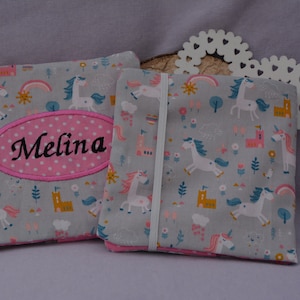 Puede incluir: Dos bolsas de tela grises con un patrón de unicornio y arcoíris. La bolsa de la izquierda tiene un óvalo rosa con el nombre "Melina" bordado.