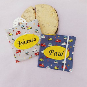 Può includere: Due quaderni rivestiti in tessuto con motivi di veicoli. Uno è grigio con un ovale giallo con "Johannes" in nero. L'altro è blu con un ovale giallo con "Paul" in nero. Entrambi hanno un elastico bianco.