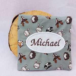 Pode incluir: Uma bolsa de tecido verde claro com um remendo oval branco que diz "Michael" em bordado marrom. A bolsa é decorada com um padrão repetido de rostos de animais marrons e brancos, incluindo veados, coelhos e ouriços.
