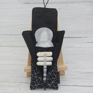 Puede incluir: Bolsa de tela negra con estampado geométrico y cremallera, que contiene productos de higiene femenina. La bolsa está abierta, revelando compresas y tampones. La solapa superior se cierra con broches y tiene un cordón negro.