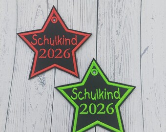 Pode incluir: Dois patches bordados em forma de estrela. Um é preto com contorno vermelho e o texto "Schulkind 2026" em vermelho. O outro é preto com contorno verde e o texto "Schulkind 2026" em verde.