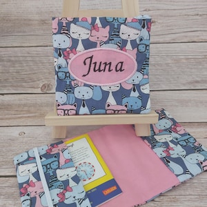 Può includere: Una copertina di libro in tessuto blu con un motivo ripetuto di gatti dei cartoni animati. La copertina ha un ovale rosa con il nome "Juna" ricamato. Sono visibili anche una copertina di libro aperta abbinata e un libro.