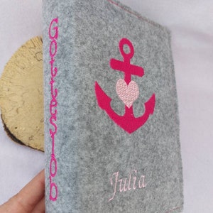 Pode incluir: Uma capa de livro cinza de feltro com um desenho de âncora e coração bordado em rosa. A capa também apresenta o nome "Julia" e a palavra "Gotteslob" em bordado rosa. Feito de material de feltro macio.