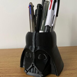 May include: A black plastic Darth Vader head desk organiser with a variety of pens and pencils inside.