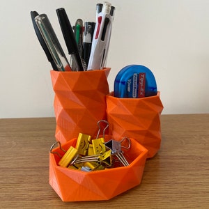 Può includere: Organizzatore da scrivania geometrico arancione con tre scomparti. Gli scomparti contengono penne, matite e graffette. Le graffette hanno facce sorridenti. L'organizzatore è realizzato in plastica stampata in 3D.