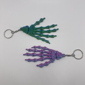 Puede incluir: Dos llaveros de mano de esqueleto, uno verde y otro morado, con anillas de llavero plateadas.