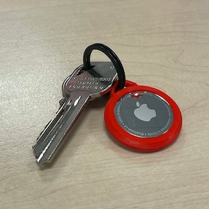 Può includere: Una chiave argentata con un portachiavi circolare rosso. Il portachiavi ha il logo Apple e il testo "Designed by Apple in California. Assembled in China. Bluetooth LE. Wideband. "
