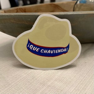 May include: A sticker featuring a light yellow cowboy hat design with a blue band. The text "¡QUE CHAVIENDA!" is printed on the blue band in white letters. The sticker has a white border and is on a neutral background.