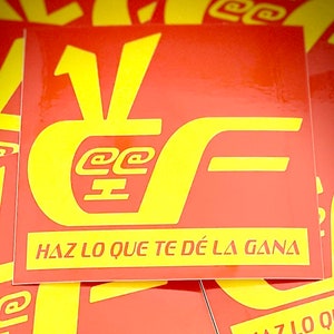 May include: A red square sticker with yellow text. The sticker features stylized letters and symbols, with the phrase "HAZ LO QUE TE DÉ LA GANA" in yellow at the bottom. The sticker has a glossy finish.