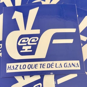 May include: A stack of blue square stickers with a white logo and text. The logo features stylized letters and the text "HAZ LO QUE TE DÉ LA GANA" in a white sans-serif font.