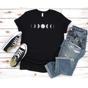 May include: Black t-shirt featuring a white graphic of the moon phases. The shirt is laid flat on a white wooden surface with a pair of black trainers and blue jeans.