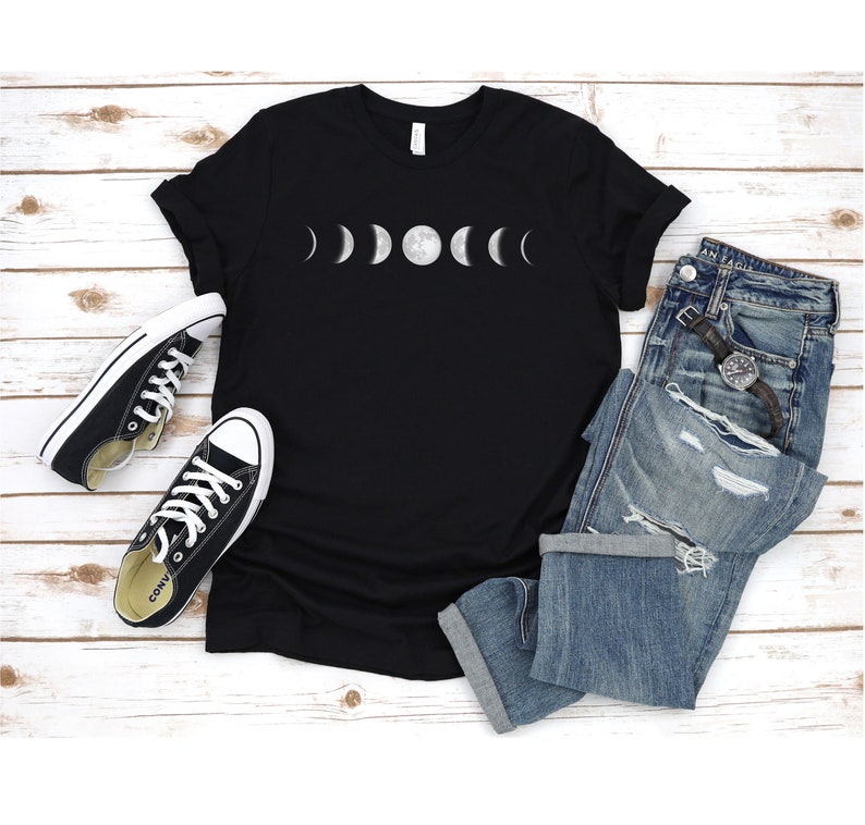 May include: Black t-shirt featuring a white graphic of the moon phases. The shirt is laid flat on a white wooden surface with a pair of black trainers and blue jeans.