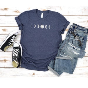 May include: A navy blue t-shirt featuring a white graphic of the moon phases. The shirt is laid flat on a white wooden surface with a pair of black canvas shoes and a pair of blue jeans.