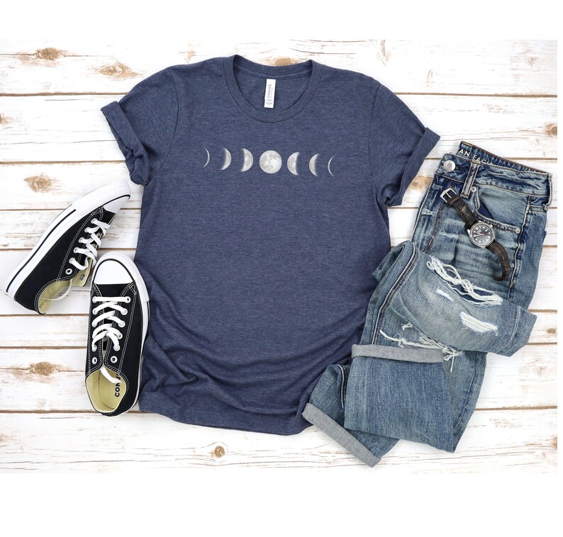May include: A navy blue t-shirt featuring a white graphic of the moon phases. The shirt is laid flat on a white wooden surface with a pair of black canvas shoes and a pair of blue jeans.
