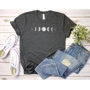 May include: A dark grey t-shirt featuring a white graphic of the moon phases. The shirt is laid out on a white wooden surface with a pair of white trainers and a pair of blue jeans.