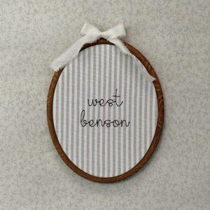 Puede incluir: Bastidor de bordado ovalado con un fondo de tela a rayas beige y blanco. El texto "west benson" está bordado en escritura negra. Una cinta blanca está atada en la parte superior.
