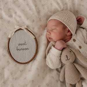 Könnte beinhalten: Ein schlafender Säugling mit einer cremefarbenen Rippmütze und einem langärmeligen Oberteil hält ein kleines beiges Strickkaninchen. Daneben liegt ein runder Holzstickrahmen mit grauen und weißen Streifen und dem Text west benson.