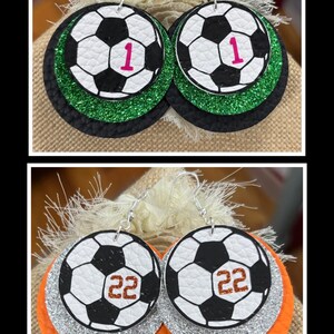 May include: Soccer ball earrings with a black and white design, featuring the number 1 in pink and the number 22 in brown. The earrings have layers of black, green, silver, and orange glitter.