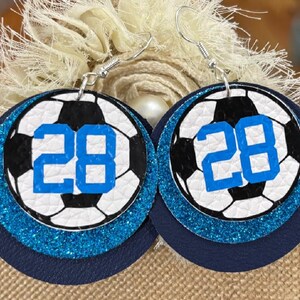 May include: A pair of round earrings featuring a soccer ball design with the number 28 in blue. The earrings have a layered effect with a navy blue base, a glittery blue middle layer, and a soccer ball print on top.