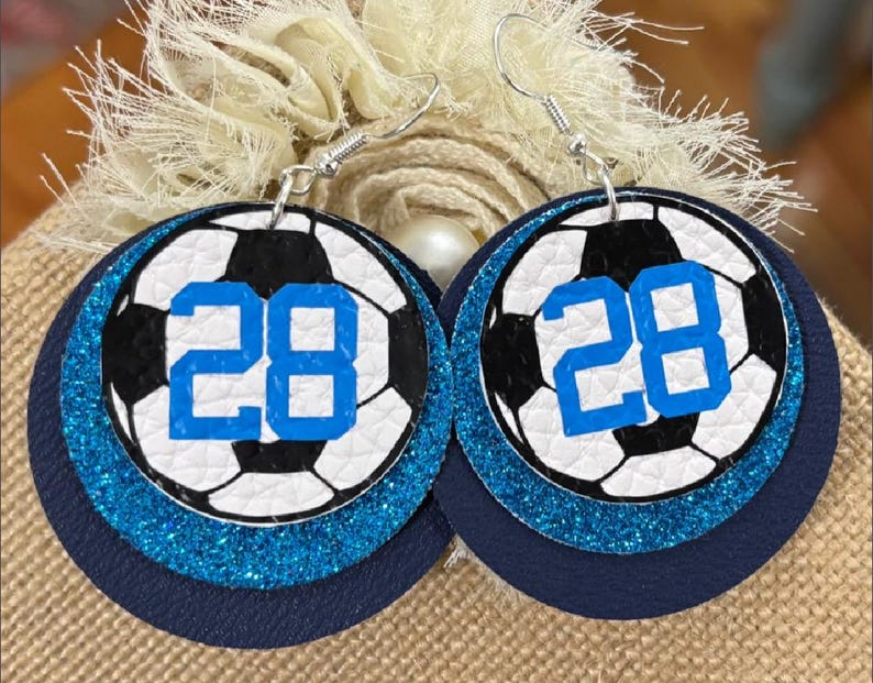 May include: A pair of round earrings featuring a soccer ball design with the number 28 in blue. The earrings have a layered effect with a navy blue base, a glittery blue middle layer, and a soccer ball print on top.