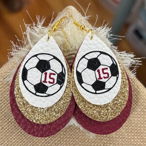 May include: Teardrop-shaped earrings with a soccer ball design. The earrings feature a white top layer with a black and white soccer ball graphic and the number 15 in red. Gold glitter and burgundy leather layers complete the design.